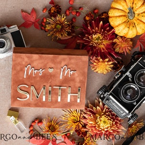 Può includere: Una scatola in pelle scamosciata marrone con il testo "Mrs. & Mr. SMITH" in lettere dorate. La scatola è circondata da decorazioni autunnali, tra cui fiori, foglie e una piccola zucca.