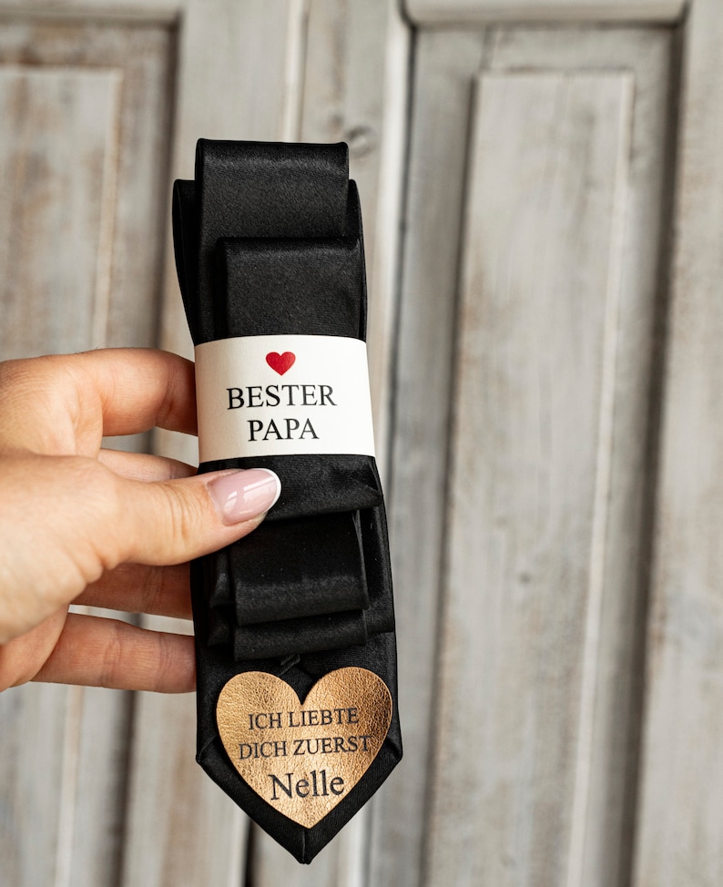 May include: Black satin tie with a white tag that says "Best Dad" with a red heart and a gold heart tag that says "I loved you first" with the name "Nelle" on it.