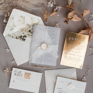 Puede incluir: Elegante juego de invitaciones de boda con un sobre forrado con flores, una invitación transparente con detalles de perlas y detalles dorados. Incluye tarjetas RSVP y una tarjeta de detalles. El conjunto está dispuesto sobre una superficie neutra con acentos florales secos.