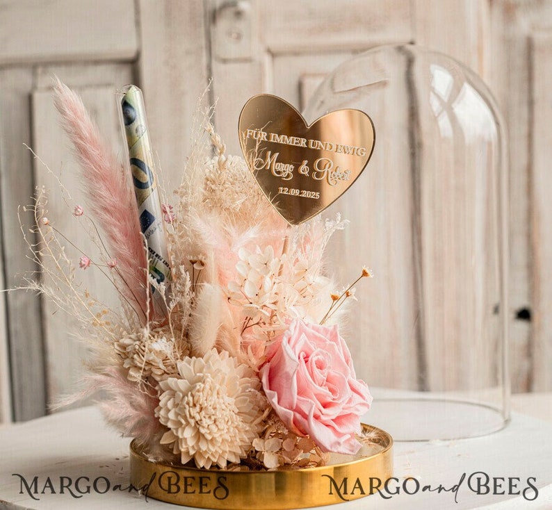 May include: A glass dome with a gold base and a pink and white preserved flower arrangement. The arrangement includes a single pink rose, dried flowers, and a gold heart-shaped plaque with the text "F&uuml;r immer und ewig Maya & Robin 12.09.2025".