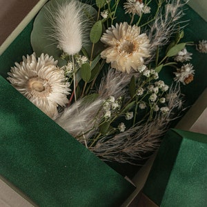 May include: A green box with a white ribbon and dried flowers inside. The flowers are white and cream colored and include baby's breath, dried daisies, and fluffy white grasses. The box has the text "MARGO and BEES" printed on the bottom.