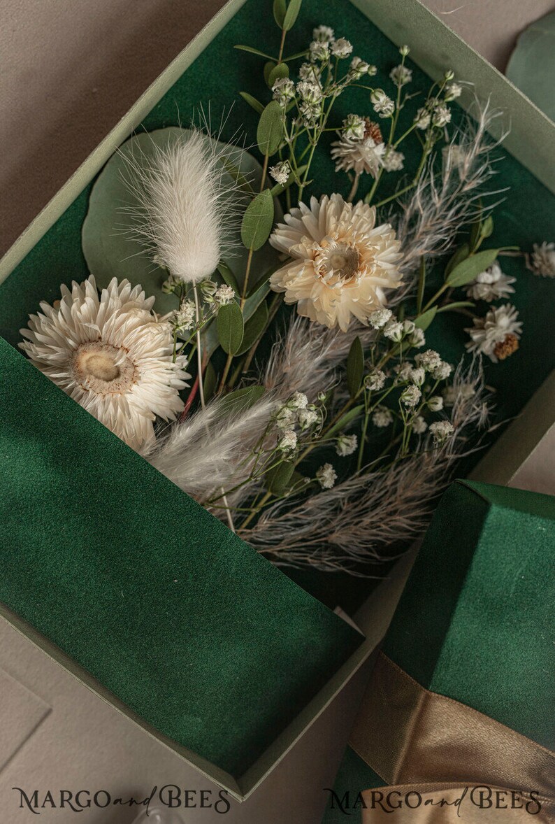 May include: A green box with a white ribbon and dried flowers inside. The flowers are white and cream colored and include baby's breath, dried daisies, and fluffy white grasses. The box has the text "MARGO and BEES" printed on the bottom.