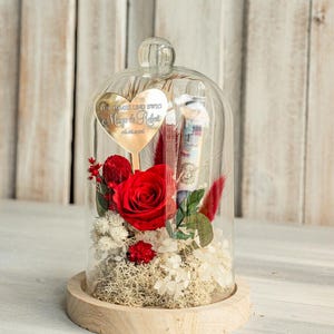 May include: A glass dome with a wooden base contains a preserved red rose, dried flowers, and a gold heart-shaped plaque with the inscription "F&uuml;r immer und ewig Maya & Robert 07.07.2023".