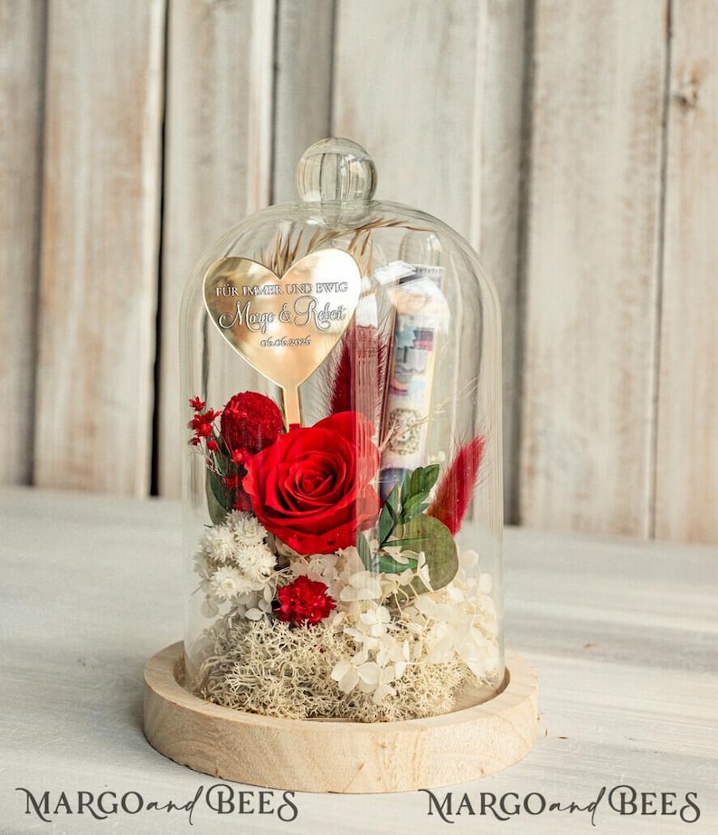 May include: A glass dome with a wooden base contains a preserved red rose, dried flowers, and a gold heart-shaped plaque with the inscription "F&uuml;r immer und ewig Maya & Robert 07.07.2023".