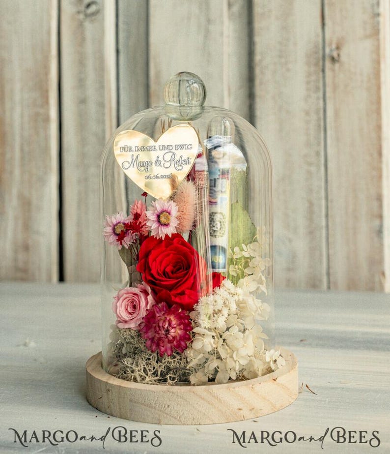 May include: A glass dome with a wooden base contains preserved flowers, including a red rose, pink roses, white hydrangeas, and other small flowers. A gold heart-shaped plaque with the text "F&uuml;r immer und ewig Maya & Robert 01.06.2019" is attached to the dome.