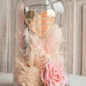 May include: A glass dome with a gold base and a gold heart-shaped plaque inside. The plaque has the text "F&uuml;r immer und ewig Margo & Robert 12.09.2025" engraved on it. The dome contains dried flowers in shades of pink and white.