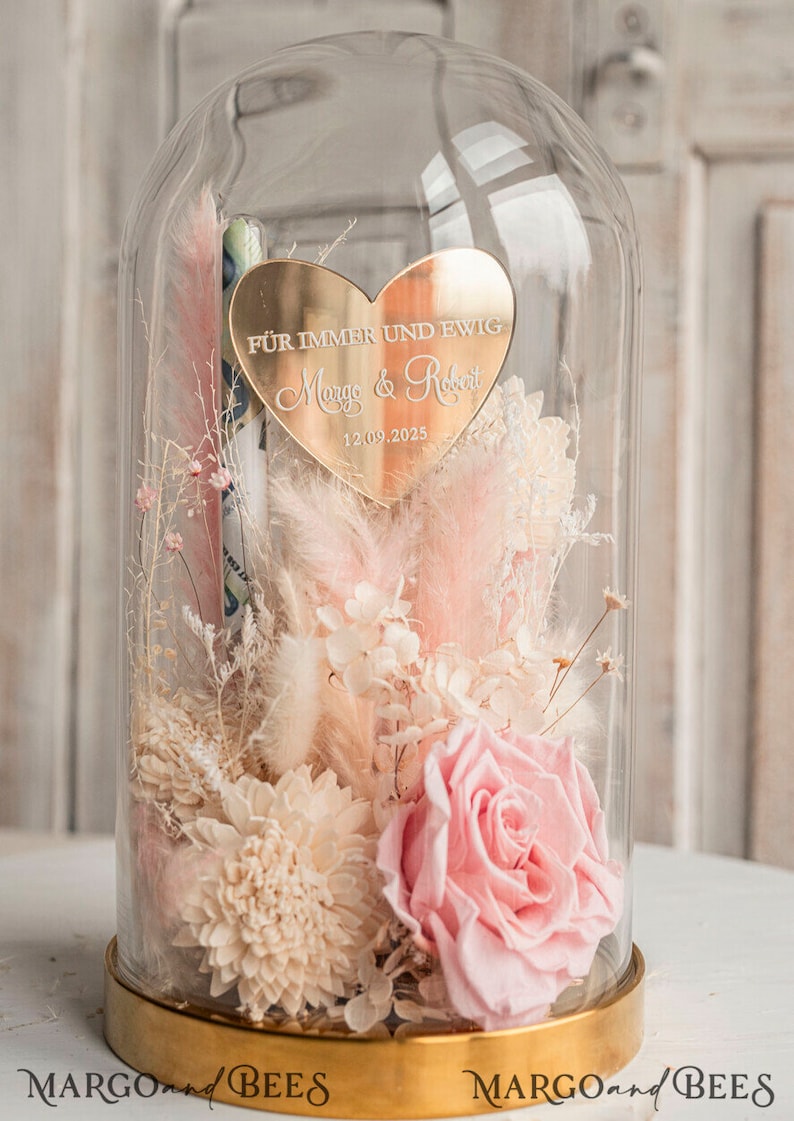 May include: A glass dome with a gold base and a gold heart-shaped plaque inside. The plaque has the text "F&uuml;r immer und ewig Margo & Robert 12.09.2025" engraved on it. The dome contains dried flowers in shades of pink and white.