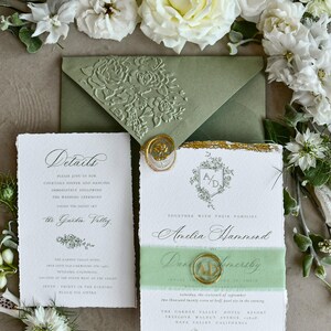 May include: A wedding invitation suite featuring a sage green envelope with a floral embossed flap, a white card with deckled edges, gold foil accents, and a gold wax seal with the initials AD, surrounded by white roses and greenery.