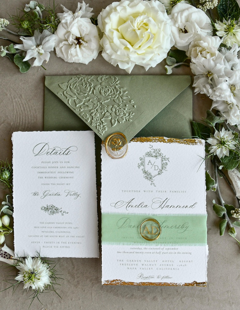 May include: A wedding invitation suite featuring a sage green envelope with a floral embossed flap, a white card with deckled edges, gold foil accents, and a gold wax seal with the initials AD, surrounded by white roses and greenery.