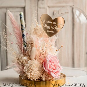 May include: A glass dome with a gold base and a pink and white dried flower arrangement. A gold heart with the text "F&uuml;r immer und ewig Margo & Robert 12.09.2025" is in the center of the arrangement.