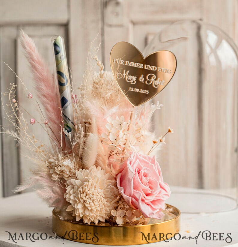 May include: A glass dome with a gold base and a pink and white dried flower arrangement. A gold heart with the text "F&uuml;r immer und ewig Margo & Robert 12.09.2025" is in the center of the arrangement.