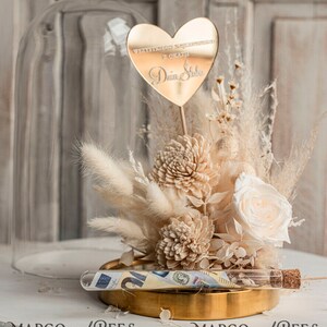 Op de afbeelding: Een glazen stolp met een gouden basis en een boeket gedroogde bloemen. De bloemen zijn wit en cr&egrave;mekleurig en omvatten pampasgras, witte rozen en gedroogde hortensia's. Een gouden hartvormige spiegel met de tekst "Wszystkiego najlepszego z okazji Dnia Slubu" is in het midden van het boeket geplaatst. Een glazen buis met een kurkstop bevat geld en is op de gouden basis geplaatst.