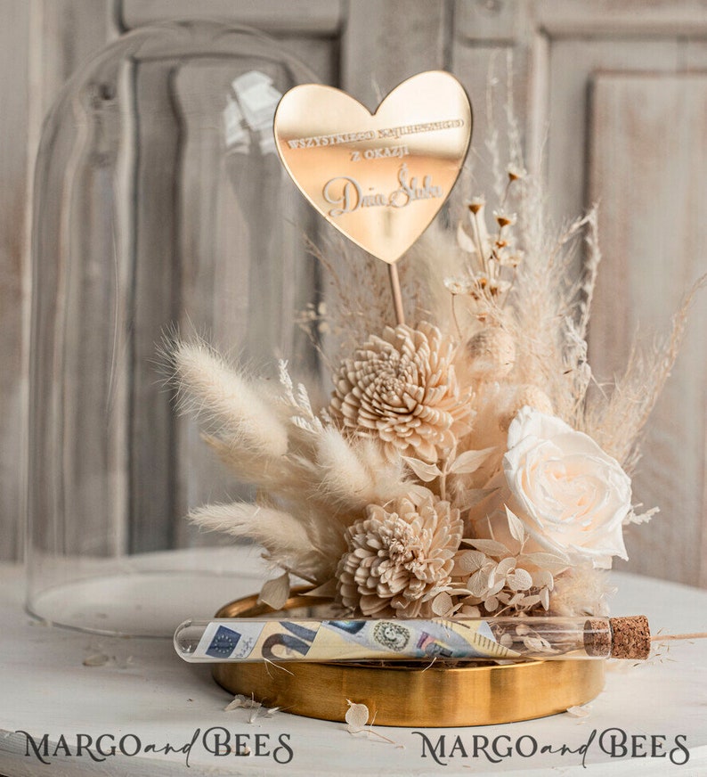 Op de afbeelding: Een glazen stolp met een gouden basis en een boeket gedroogde bloemen. De bloemen zijn wit en cr&egrave;mekleurig en omvatten pampasgras, witte rozen en gedroogde hortensia's. Een gouden hartvormige spiegel met de tekst "Wszystkiego najlepszego z okazji Dnia Slubu" is in het midden van het boeket geplaatst. Een glazen buis met een kurkstop bevat geld en is op de gouden basis geplaatst.