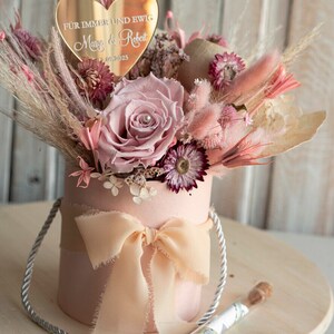 May include: A pink box with a white ribbon and a bouquet of dried flowers. The flowers are pink, white, and beige. A gold heart-shaped plaque with the text "F&uuml;r immer und ewig" and the names "Mary & Robert" and the date "21.09.2025" is placed on top of the bouquet.