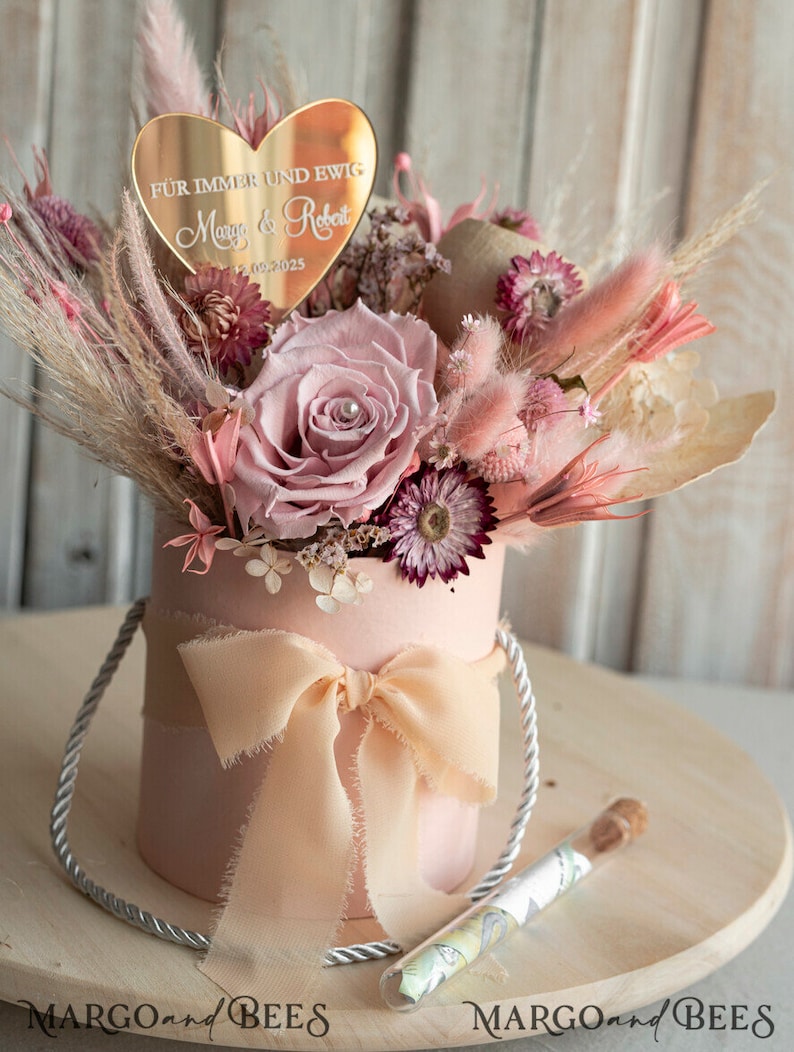 May include: A pink box with a white ribbon and a bouquet of dried flowers. The flowers are pink, white, and beige. A gold heart-shaped plaque with the text "F&uuml;r immer und ewig" and the names "Mary & Robert" and the date "21.09.2025" is placed on top of the bouquet.