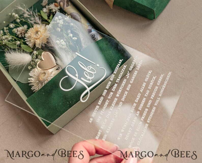 May include: A clear acrylic box with a green velvet interior and a gold heart charm. The box contains dried flowers and a clear acrylic card with the text "Lieb!" and a wedding message in German.