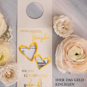 May include: A white card with gold lettering that says "WEINETIKETT - GELDUMSCHLAG" and "MIT DEN BESTEN W&Uuml;NSCHEN" with two gold heart cutouts. The card has a slot for money and the text "F&Uuml;R EURS GEMEINSAME ZUKUNFT" and "HIER DAS GELD EINLEGEN". The card is decorated with flowers.