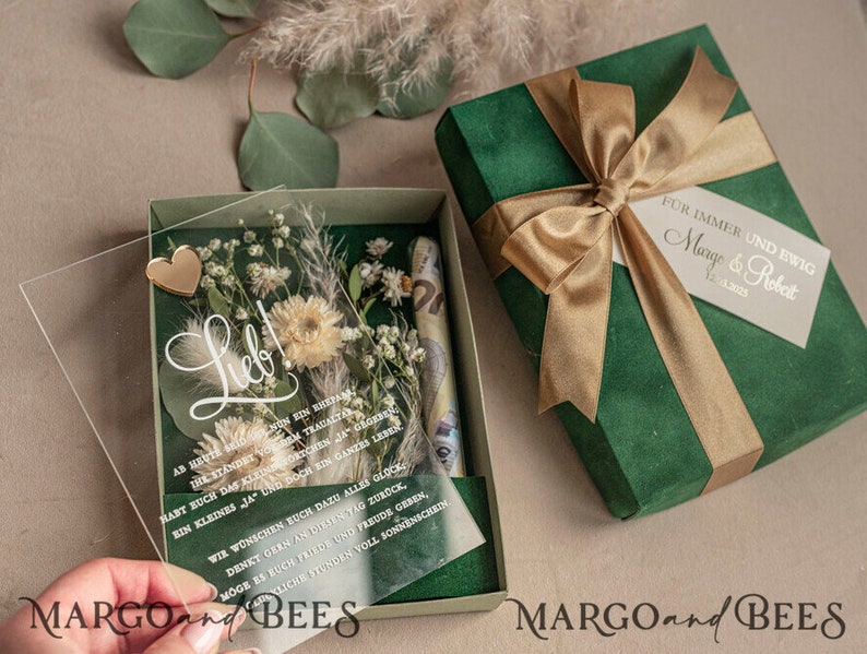 May include: A green gift box with a gold ribbon and a white tag that says "For immer und ewig Margo & Robert 1. Juni 2022". The box is open and inside is a clear acrylic box with dried flowers, a gold heart, and a message that says "Lieb!"  The message continues with a poem in German.