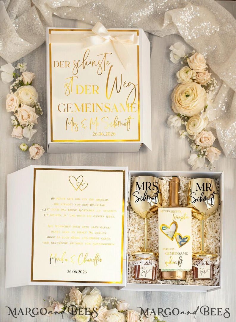 May include: A white gift box with a gold inscription that reads "Der sch&ouml;nste Weg ist der gemeinsame Mrs & Mr Schmidt 26.06.2026". The box contains two gold-rimmed wine glasses, a bottle of wine, and two small jars of jam. The glasses are engraved with "Mrs Schmidt" and "Mr Schmidt".
