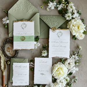 May include: A wedding stationery set featuring sage green envelopes with embossed floral flaps, white paper cards with deckled edges and gold leaf accents, a gold wax seal, and a gold quill pen, surrounded by white flowers and greenery.