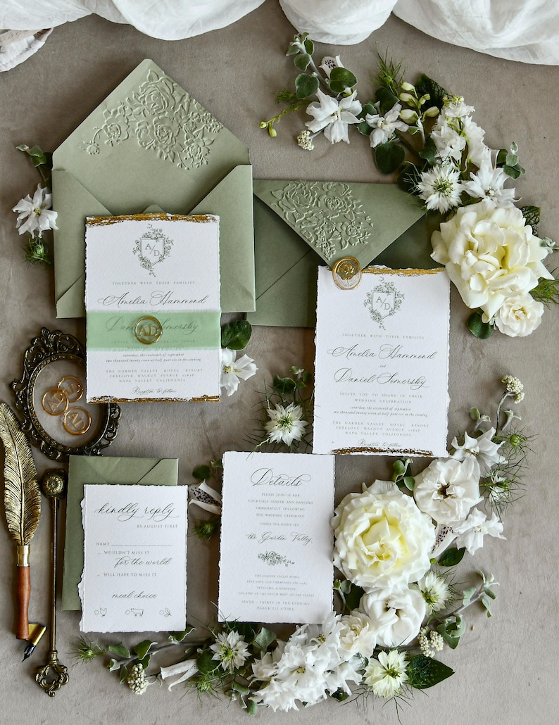 May include: A wedding stationery set featuring sage green envelopes with embossed floral flaps, white paper cards with deckled edges and gold leaf accents, a gold wax seal, and a gold quill pen, surrounded by white flowers and greenery.