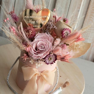 May include: A pink box with a bow and a bouquet of dried flowers. The flowers are pink, white, and brown. A gold heart-shaped plaque with the text "F&uuml;r immer und ewig Margo & Robert 12.09.2025" is placed on top of the bouquet.