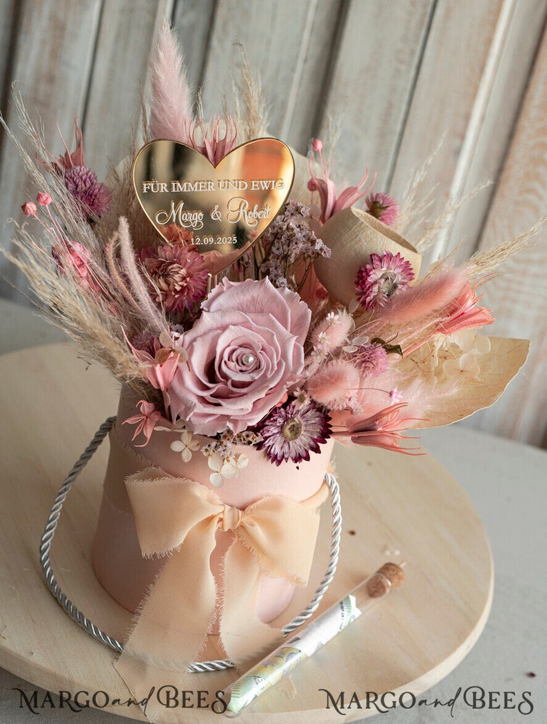 May include: A pink box with a bow and a bouquet of dried flowers. The flowers are pink, white, and brown. A gold heart-shaped plaque with the text "F&uuml;r immer und ewig Margo & Robert 12.09.2025" is placed on top of the bouquet.