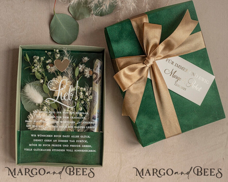 May include: A green gift box with a gold ribbon and a white tag that says "For always and forever Margo & Robert 12.01.2025". The box is open and inside is a clear box with dried flowers and a heart-shaped charm. The text "Lieb!" is written on the clear box. The box is labeled "MARGO and BEES".