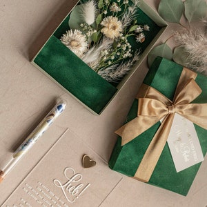 May include: A green velvet gift box with a gold ribbon and a white tag that says "Forever and Ever, Margo & Robert 12.2022". The box is sitting on a beige surface next to a clear acrylic card with the word "Lieb!" written in white cursive. The card also has a small gold heart charm attached to it. There is a clear glass tube with money inside.