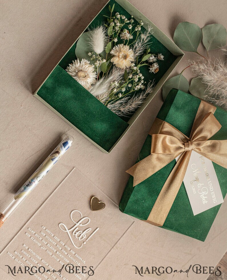 May include: A green velvet gift box with a gold ribbon and a white tag that says "Forever and Ever, Margo & Robert 12.2022". The box is sitting on a beige surface next to a clear acrylic card with the word "Lieb!" written in white cursive. The card also has a small gold heart charm attached to it. There is a clear glass tube with money inside.