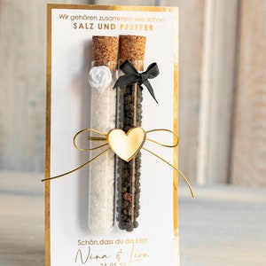May include: A white card with gold trim features a wedding favor with two glass test tubes filled with salt and pepper. The test tubes are tied together with a gold ribbon and a gold heart charm. The card reads "Wir geh&ouml;ren zusammen wie schon. SALZ UND PFEFFER" and "Sch&ouml;n, dass du da bist! Nina & Leon 25.05.25" with the text "MARGOand BEES" at the bottom.