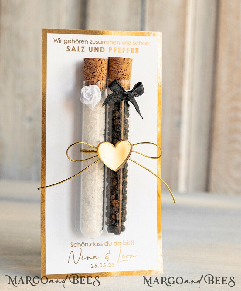 May include: A white card with gold trim features a wedding favor with two glass test tubes filled with salt and pepper. The test tubes are tied together with a gold ribbon and a gold heart charm. The card reads "Wir geh&ouml;ren zusammen wie schon. SALZ UND PFEFFER" and "Sch&ouml;n, dass du da bist! Nina & Leon 25.05.25" with the text "MARGOand BEES" at the bottom.
