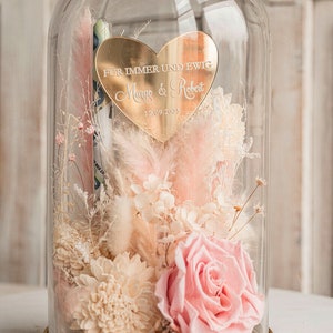 May include: A glass dome with a gold base and a heart-shaped plaque inside. The plaque reads "F&uuml;r immer und ewig Margo & Robert 12.09.2025". The dome contains dried flowers in shades of pink and white.
