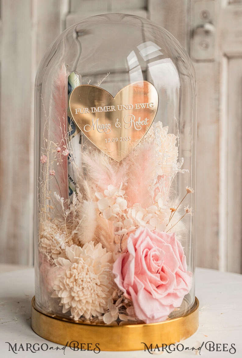 May include: A glass dome with a gold base and a heart-shaped plaque inside. The plaque reads "F&uuml;r immer und ewig Margo & Robert 12.09.2025". The dome contains dried flowers in shades of pink and white.