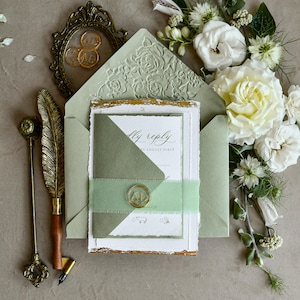 May include: A sage green envelope with a floral embossed flap, containing a white invitation card with deckled edges and gold leaf accents. A smaller sage green card is secured with a light green ribbon and a gold wax seal featuring the letters AD.