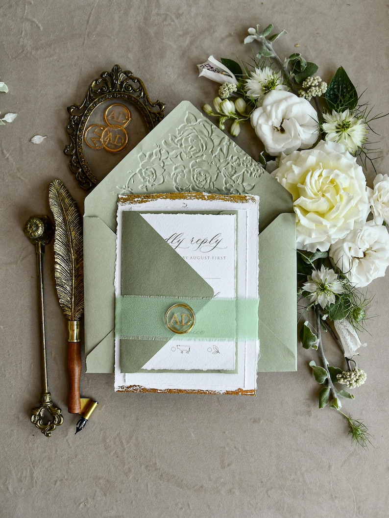 May include: A sage green envelope with a floral embossed flap, containing a white invitation card with deckled edges and gold leaf accents. A smaller sage green card is secured with a light green ribbon and a gold wax seal featuring the letters AD.