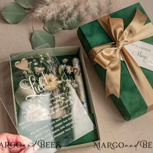 May include: A green velvet gift box with a gold ribbon and a clear acrylic box with dried flowers and a gold heart. The acrylic box has a handwritten message that says "Lieb!" and a text that reads "Habt euch das Wortchen ja doch ein ganzes Leben lang gegeben."