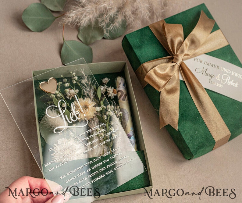 May include: A green velvet gift box with a gold ribbon and a clear acrylic box with dried flowers and a gold heart. The acrylic box has a handwritten message that says "Lieb!" and a text that reads "Habt euch das Wortchen ja doch ein ganzes Leben lang gegeben."
