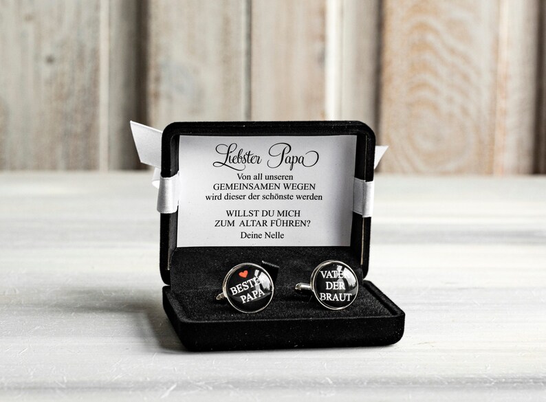 May include: A black velvet box with a white card that reads "Liebster Papa" and a message about a wedding. The box contains two silver cufflinks with black enamel and white text. One cufflink says "Beste Papa" with a red heart and the other says "Vate der Braut".