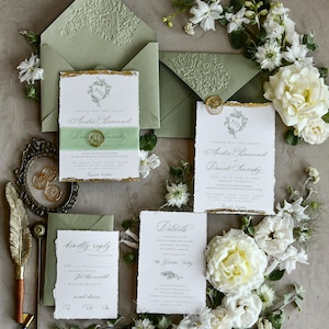 May include: A wedding stationery set featuring white paper cards with deckled edges and gold foil accents, sage green envelopes with embossed floral flaps, a green vellum belly band with a gold wax seal, and a gold quill pen with a metal frame.