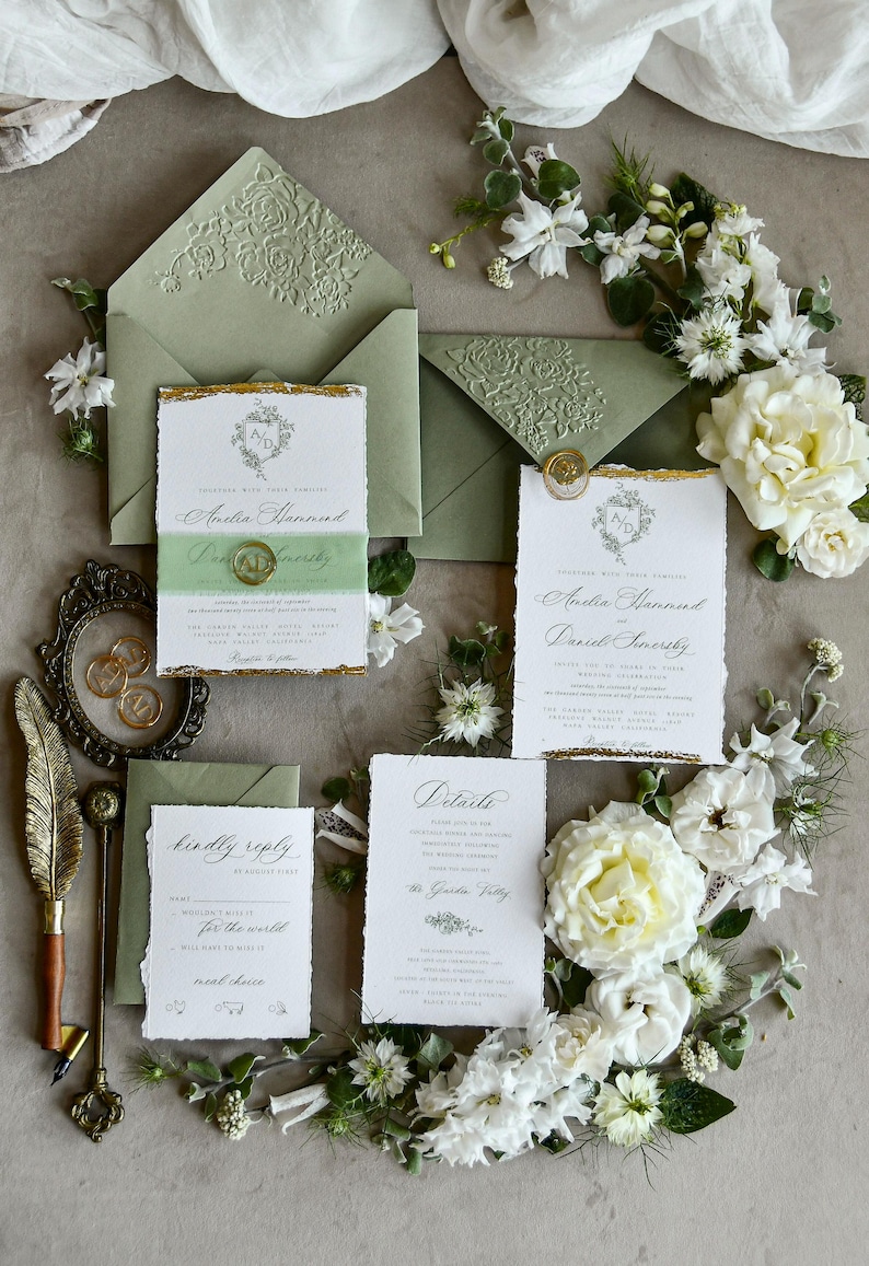 May include: A wedding stationery set featuring white paper cards with deckled edges and gold foil accents, sage green envelopes with embossed floral flaps, a green vellum belly band with a gold wax seal, and a gold quill pen with a metal frame.