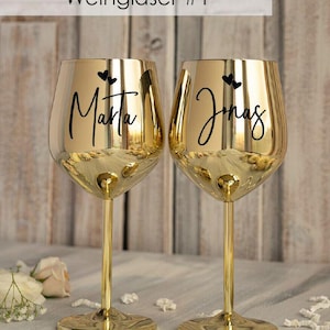 May include: Two gold-colored wine glasses with black lettering. The glasses are engraved with the names "Marta" and "Jonas" and two small hearts.