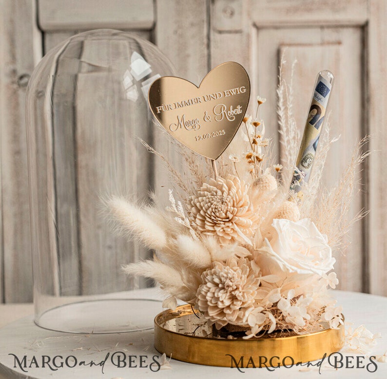 Op de afbeelding: Een glazen stolp met een gouden basis en een boeket gedroogde bloemen erin. De bloemen zijn wit en cr&egrave;mekleurig en omvatten rozen, pampasgras en andere gedroogde bloemen. Een gouden hartvormige spiegel met de tekst "FOR IMMER UND EWIG" en de namen "Maya & Robert" en de datum "12.09.2025" is in het boeket geplaatst. Een doorzichtig reageerbuisje met geld erin is ook in het boeket geplaatst.