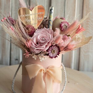 May include: A pink gift box with a white ribbon and a bouquet of dried flowers. The flowers are pink, white, and purple. A heart-shaped mirror with the text "F&uuml;r immer und ewig Margo & Robert 09.2025" is on top of the flowers.