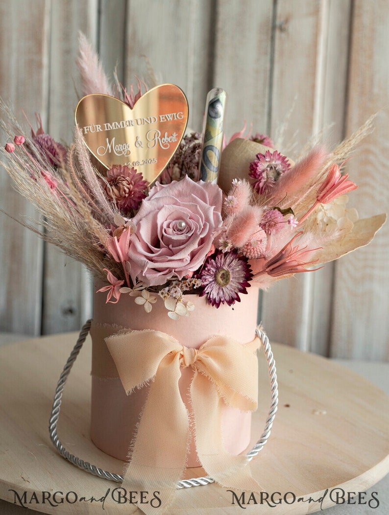 May include: A pink gift box with a white ribbon and a bouquet of dried flowers. The flowers are pink, white, and purple. A heart-shaped mirror with the text "F&uuml;r immer und ewig Margo & Robert 09.2025" is on top of the flowers.