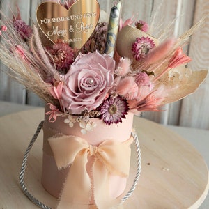 May include: A pink box with a peach-coloured ribbon and a bouquet of dried flowers. The bouquet includes a single pink rose, pink and white pampas grass, and other dried flowers. A gold heart-shaped sign with the text "F&uuml;r immer und ewig Margo & Robert 12.09.2025" is placed on top of the bouquet.