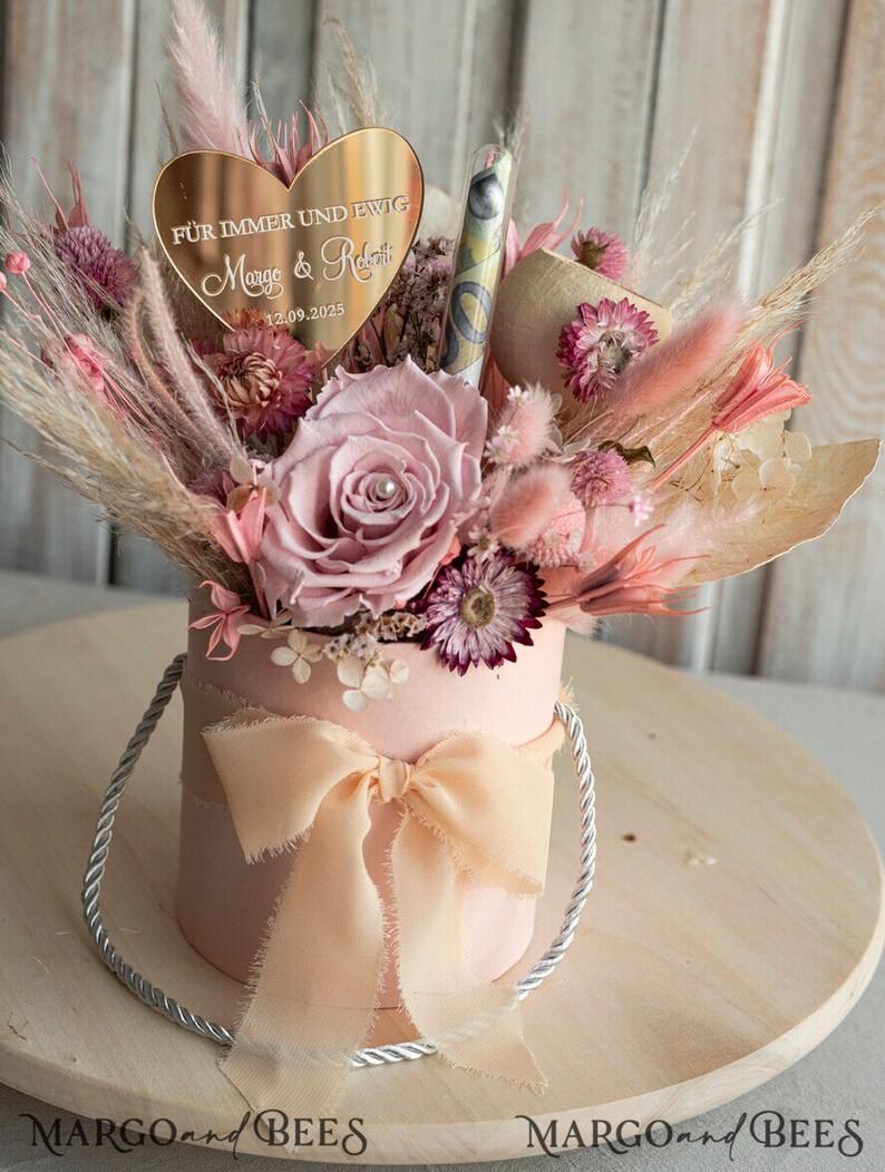 May include: A pink box with a peach-coloured ribbon and a bouquet of dried flowers. The bouquet includes a single pink rose, pink and white pampas grass, and other dried flowers. A gold heart-shaped sign with the text "F&uuml;r immer und ewig Margo & Robert 12.09.2025" is placed on top of the bouquet.
