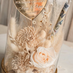 Op de afbeelding: Een glazen stolp met een gouden basis en een hartvormig bord met de tekst "F&Uuml;R IMMER UND EWIG Margo & Robert 13.08.2025". In de stolp bevinden zich gedroogde bloemen in cr&egrave;me- en wit tinten, waaronder een enkele perzikroos.