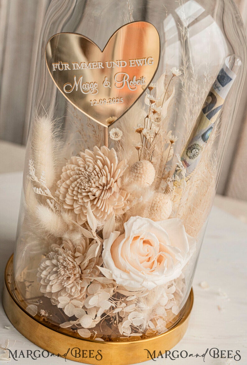 Op de afbeelding: Een glazen stolp met een gouden basis en een hartvormig bord met de tekst "F&Uuml;R IMMER UND EWIG Margo & Robert 13.08.2025". In de stolp bevinden zich gedroogde bloemen in cr&egrave;me- en wit tinten, waaronder een enkele perzikroos.