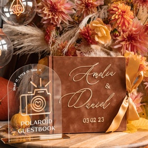 Könnte beinhalten: Ein braunes Samt-Gästebuch mit goldenen Lettern, auf dem "Amelia & Daniel 03 02 23" steht. Ein klares Acrylschild mit goldenen Lettern lautet "Snap it. Stick it. Sign it." und zeigt ein goldenes Kamerasymbol. Das Schild steht vor dem Gästebuch und befindet sich auf einer Holzoberfläche.
